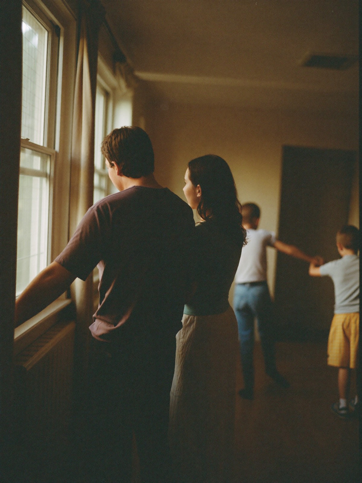 Two parents standing at the window of their home in late afternoon light, their children playing behind them.
