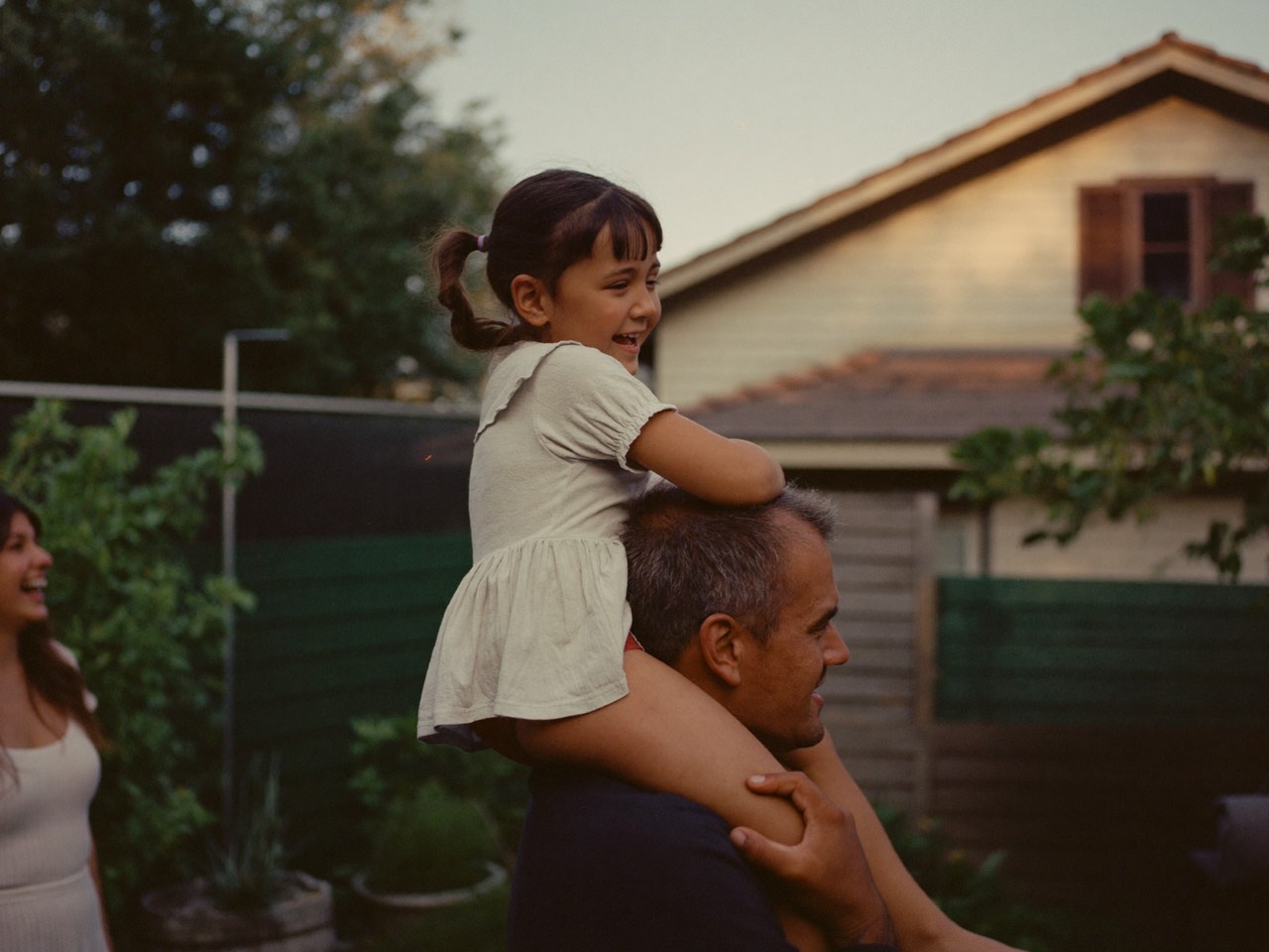 A father carrying his young daughter on his shoulders in the back garden, her mother laughing nearby.