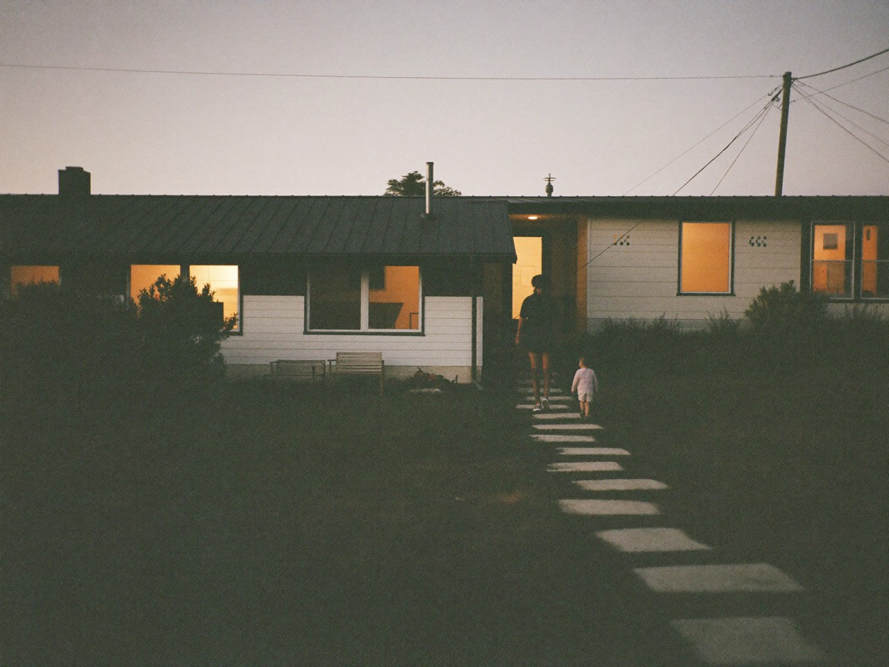 A family home at dusk with warm light in the windows, a parent and child walking up the front path.