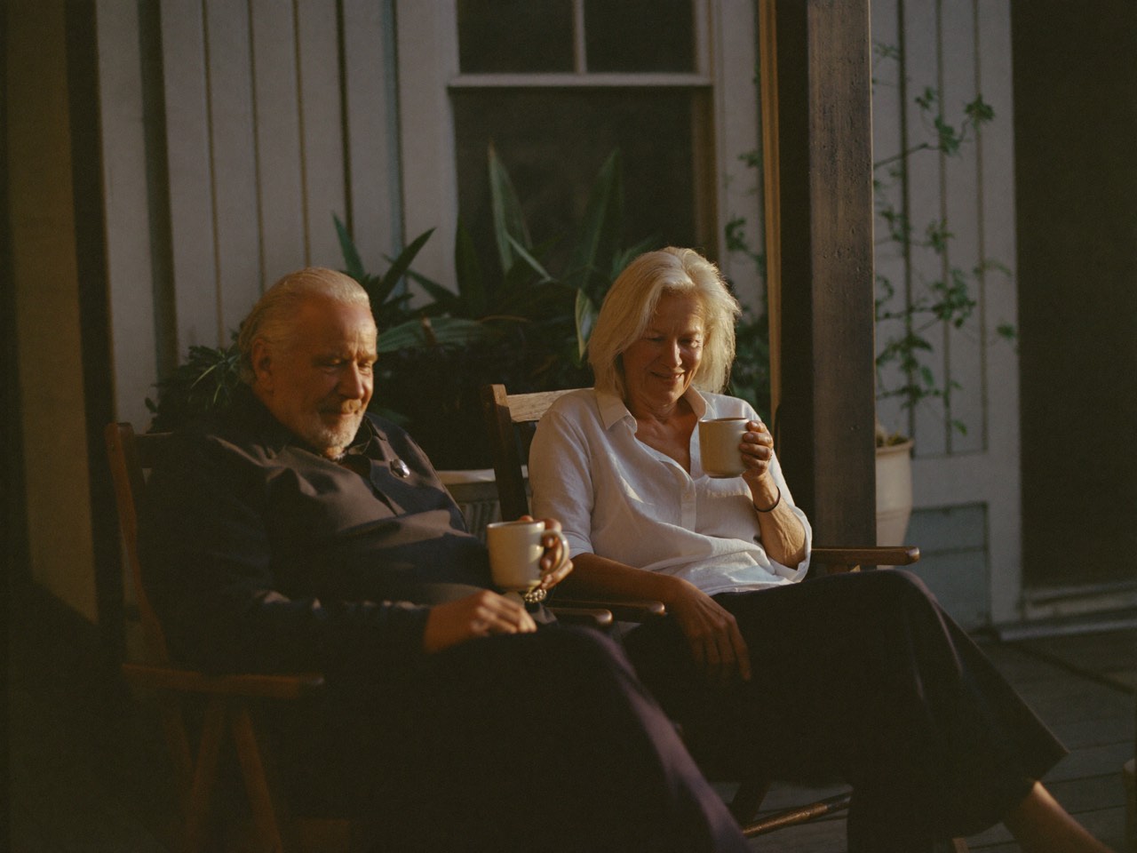 A couple in their sixties sitting together on their back porch at golden hour, holding mugs of coffee.